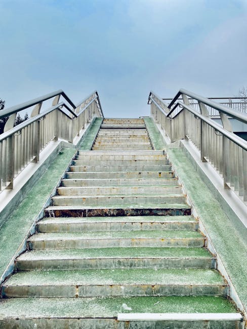 A set of outdoor metal stairs with black rubber treads marked by a yellow central line, leading from street level to an elevated pedestrian walkway or bridge. The stairs are equipped with silver metal