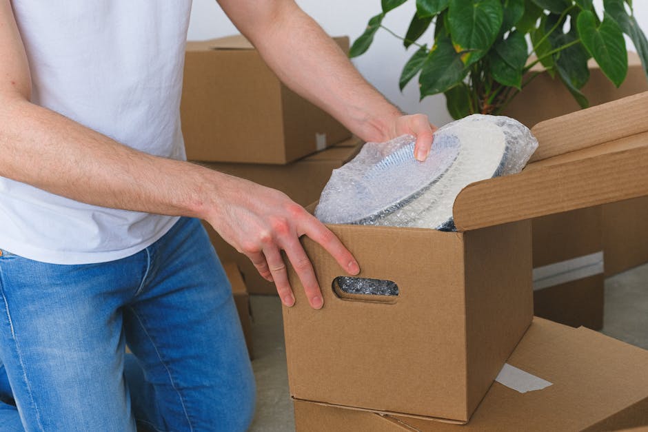 A large, wrapped antique porcelain figurine is positioned upright on a padded surface inside a living room, prepared for a house removal service. The figurine is carefully covered in transparent bubbl