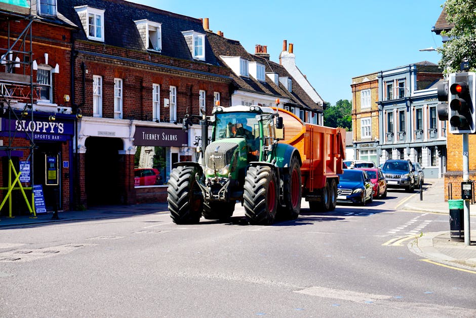 A large green tractor with an orange trailer attached is parked on a city street, surrounded by cars and buildings with mixed architectural styles. The tractor is positioned near the curb in an urban area, possibly involved in a home relocation or moving process. In the background, there are brick and painted façades of shops and residences, including a sports bar called Sloppy's and a salon named Tierney Salons. Nearby, a traffic light is visible on the corner, and a green waste bin is placed by the sidewalk. Bright daylight illuminates the scene, with the tractor seemingly preparing for furniture transport or loading activities related to house removals, reflecting typical moving logistics handled by services like Removal Company Merton.