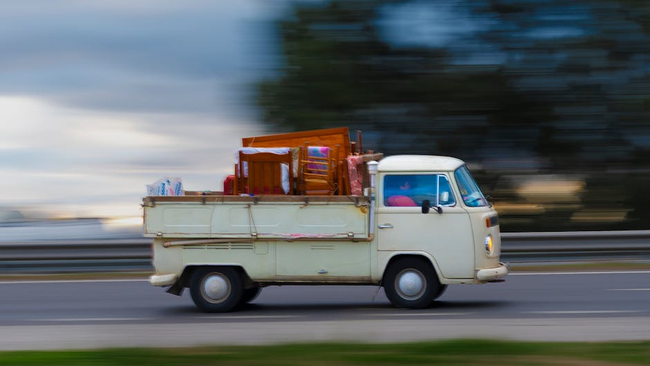 An old white van with a boxy shape is shown moving along an open road during daytime, loaded with various household furniture and items. The cargo area contains wooden chairs, a small wooden table, and several cardboard boxes wrapped in plastic, indicating packing for a home relocation. The items are secured on the flatbed with straps and are arranged to optimize space, reflecting typical furniture transport during packing and moving processes. The background features a blurred landscape with trees and a cloudy sky, emphasizing the van’s motion. The vehicle is operated by Removal Company Merton, a professional removals service, providing house moving support including loading and transportation of household possessions. The scene highlights key aspects of furniture transport and the logistical aspects of a home relocation.